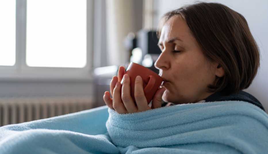 Una mujer enferma, arropada con una manta azul, se toma un té