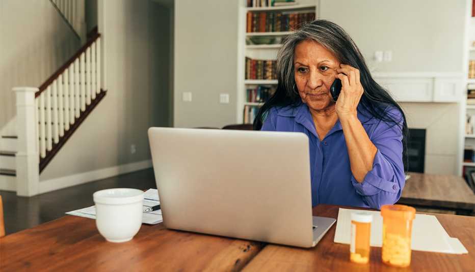Una mujer sentada frente a una computadora mientras hace una llamada y sobre la mesa tiene medicinas recetadas