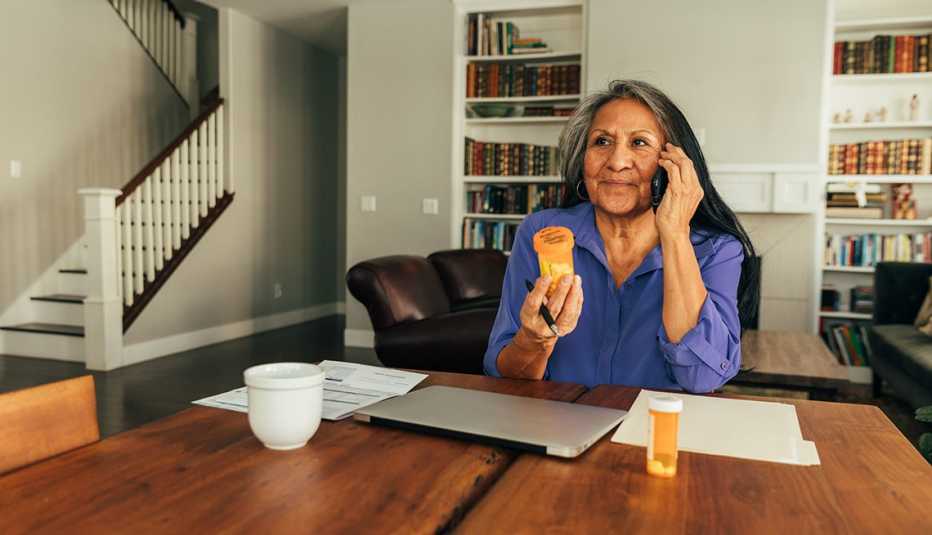 Una mujer sentada en su cocina conversa por teléfono mientras tiene un pote de pastillas en su mano
