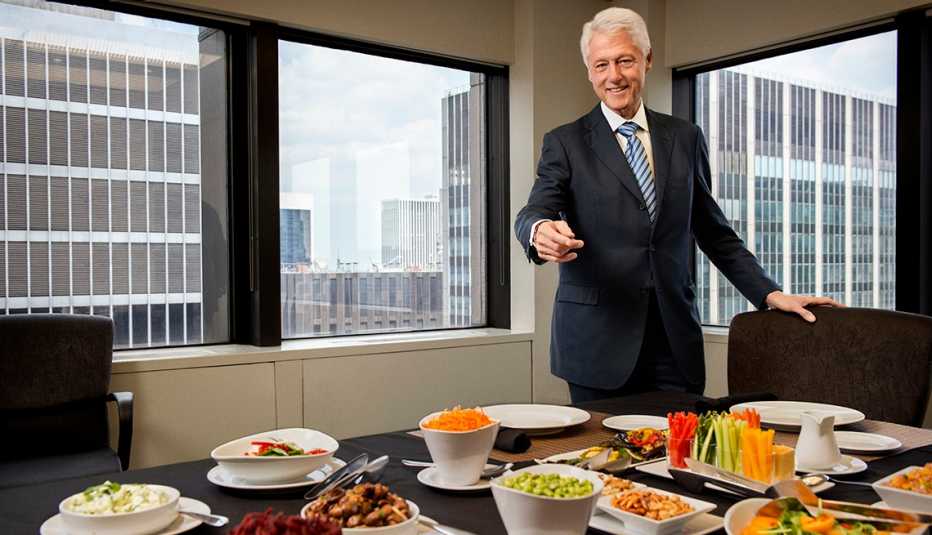 Bill Clinton sonríe y apunta hacia la mesa con platos de vegetales. 