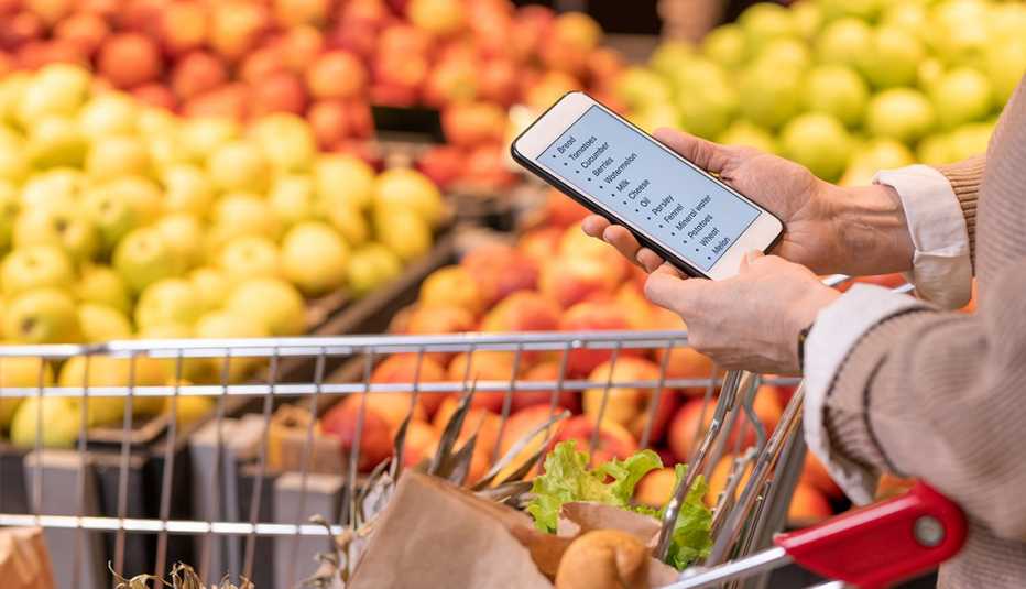 Manos de una mujer que revisa la lista de compras en su teléfono frente a la sección de frutas de un supermercado