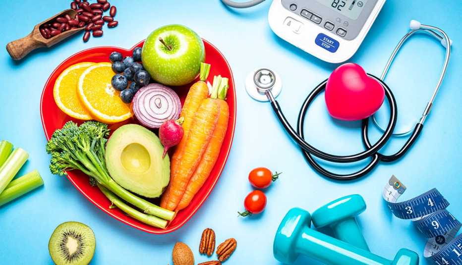 Plato en forma de corazón con frutas y vegetales, alimentos nutritivos, un estetoscopio, un corazón de goma, cinta métrica y unas mancuernas sobre un fondo azul