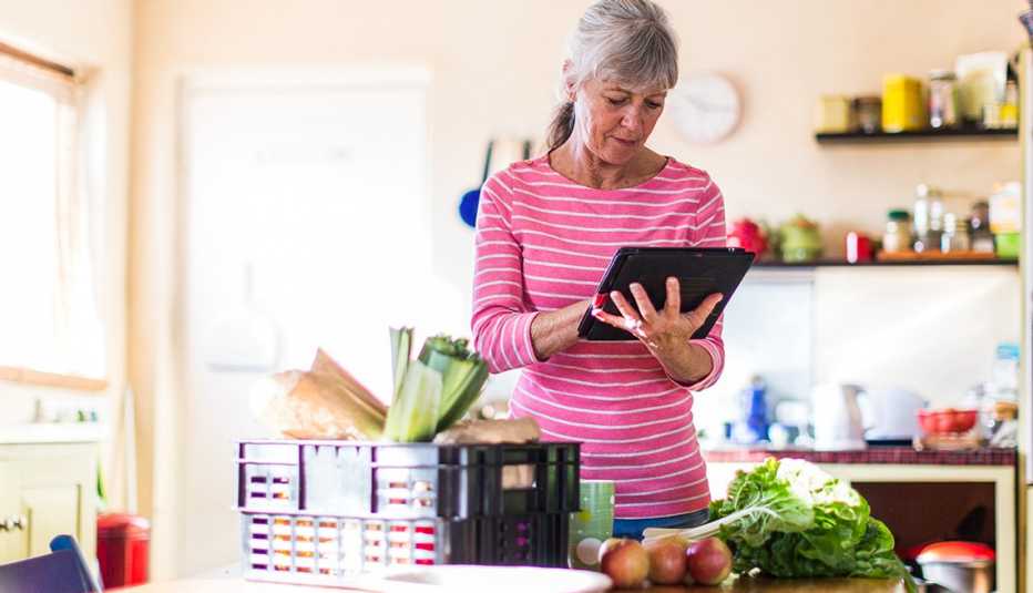 Una mujer mira su tableta parada en su cocina y alimentos sobre la mesa