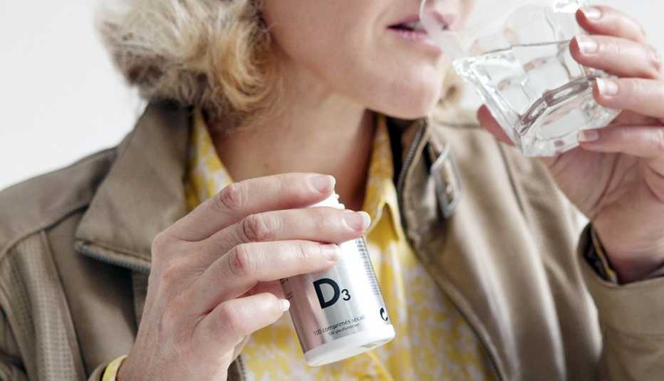 Una mujer sostiene un frasco de vitamina D en una mano mientras toma agua