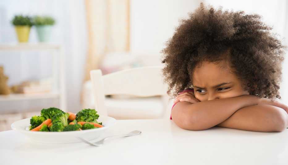Una niña con sus brazos cruzados mira de reojo y molesta un plato de brócoli y zanahorias