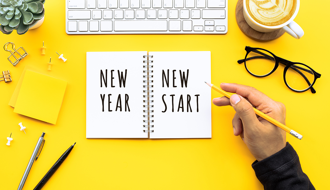 Una persona con un lápiz en mano y una libreta que dice nuevo año, nuevo comienzo en inglés. También se ve un teclado, útiles de oficina, unos espejuelos y una taza de café sobre un escritorio amarillo