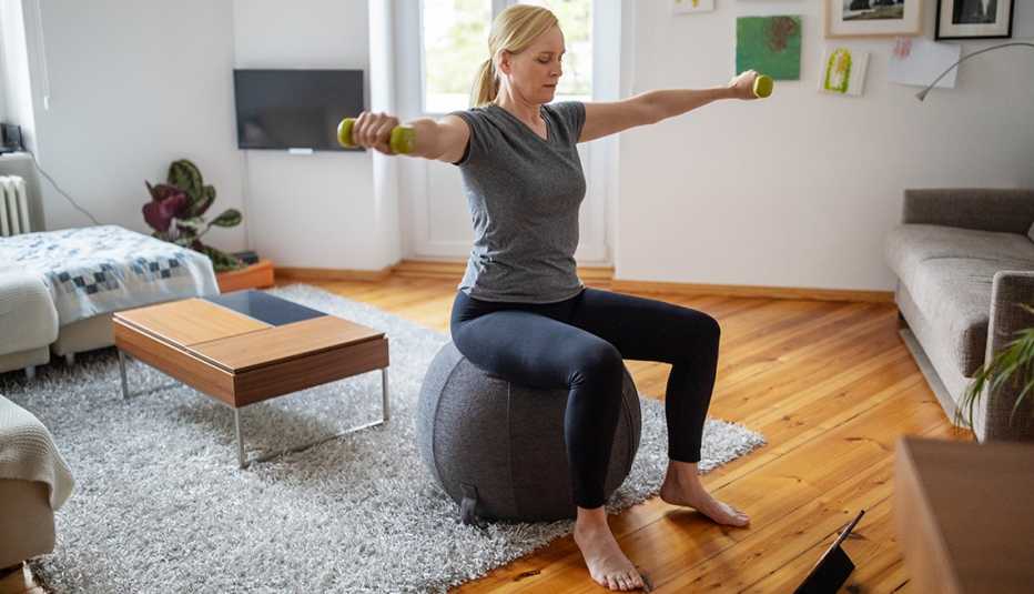 Una mujer sentada en una bola de balance levanta sus brazos hacia los lados con unas pesas livianas