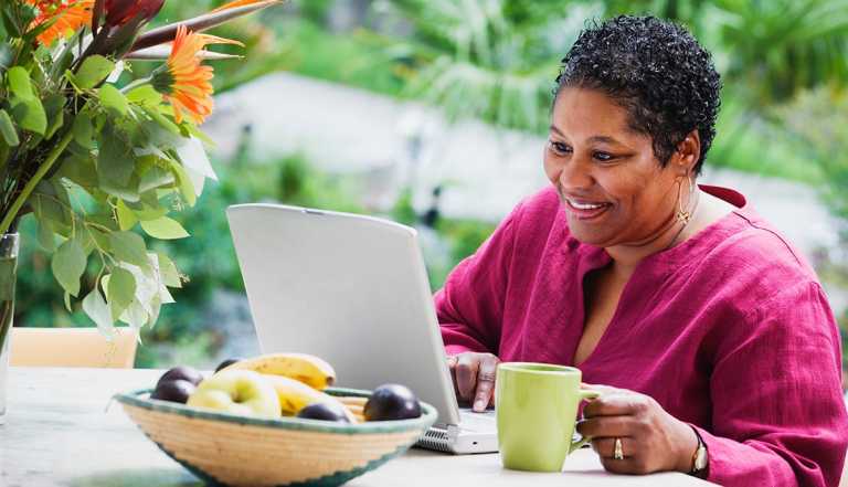 una mujer con una camisa morada sostiene su café y revisa su computadora portátil