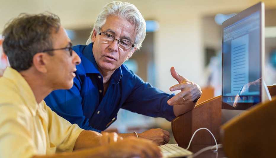 Hombre adulto aconsejando a otro que está sentado frente a un computador - Asesor de cambio de carrera 