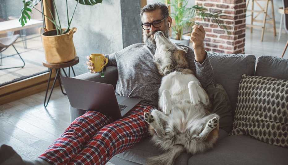 Hombre en pijama sentado en el sofá de su casa con un computador en las piernas y su perro al lado