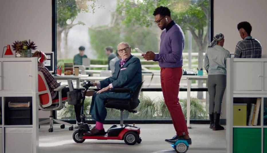 Toma de pantalla de un comercial que muestra una persona mayor en una silla eléctrica y un hombre joven en un scooter