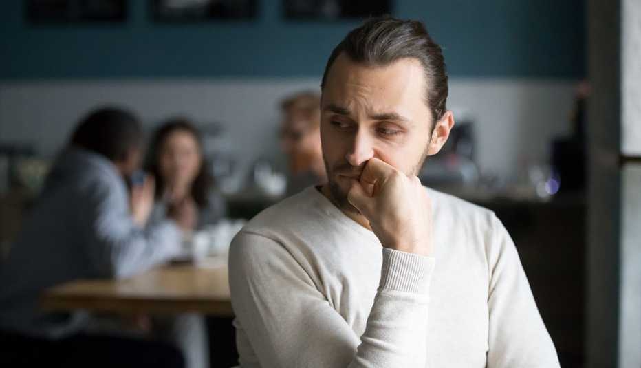 Hombre sentado en una cafetería mientras se lleva la mano a la boca y mantiene su mirada hacia abajo. 