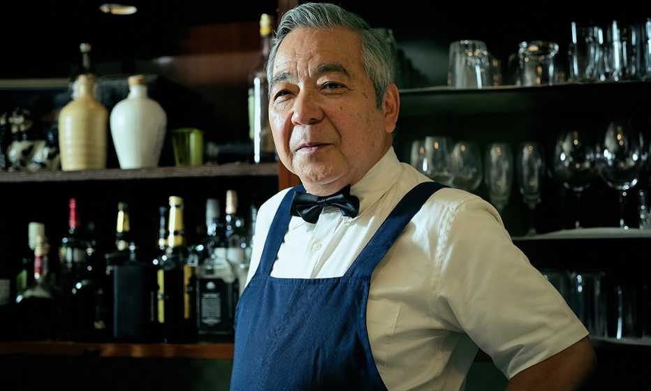 Un barman con corbata en lazo y delantal sonríe mientras está parado frente a botellas en estantes