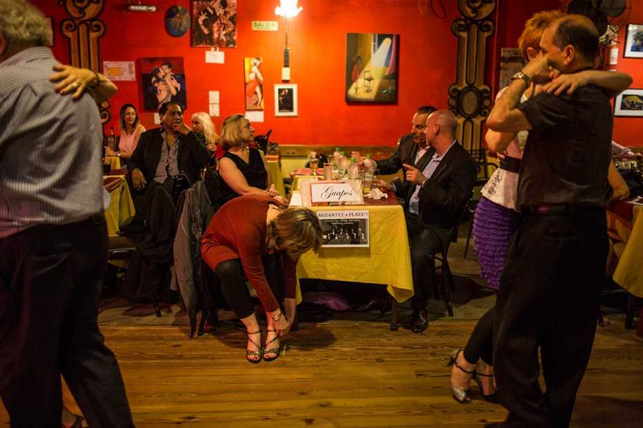 Kathleen Currie se amarra los zapatos en una mesa mientras su profesor Louis Blanco y sus compañeros de clases de tango en Little Rock, Arkansas, conversan en una mesa de una milonga en Buenos Aires. 