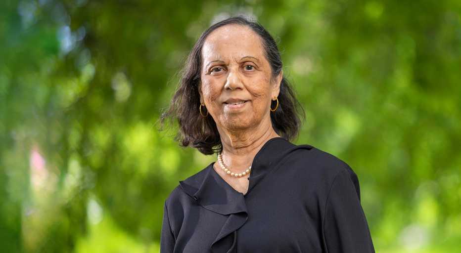 An older adult standing outdoors against a green, blurred natural background, wearing a dark top and pearl necklace.
