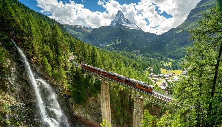 Scenic Swiss mountain train traveling across a bridge above a waterfall and lush green valley, surrounded by Alpine forests and towering peaks