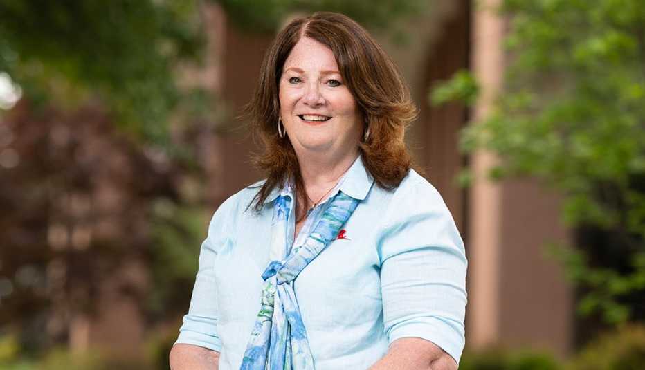 Outdoor portrait of Kathleen O’Leary seated in a garden-like setting with greenery in the background, wearing a light-colored top and patterned scarf