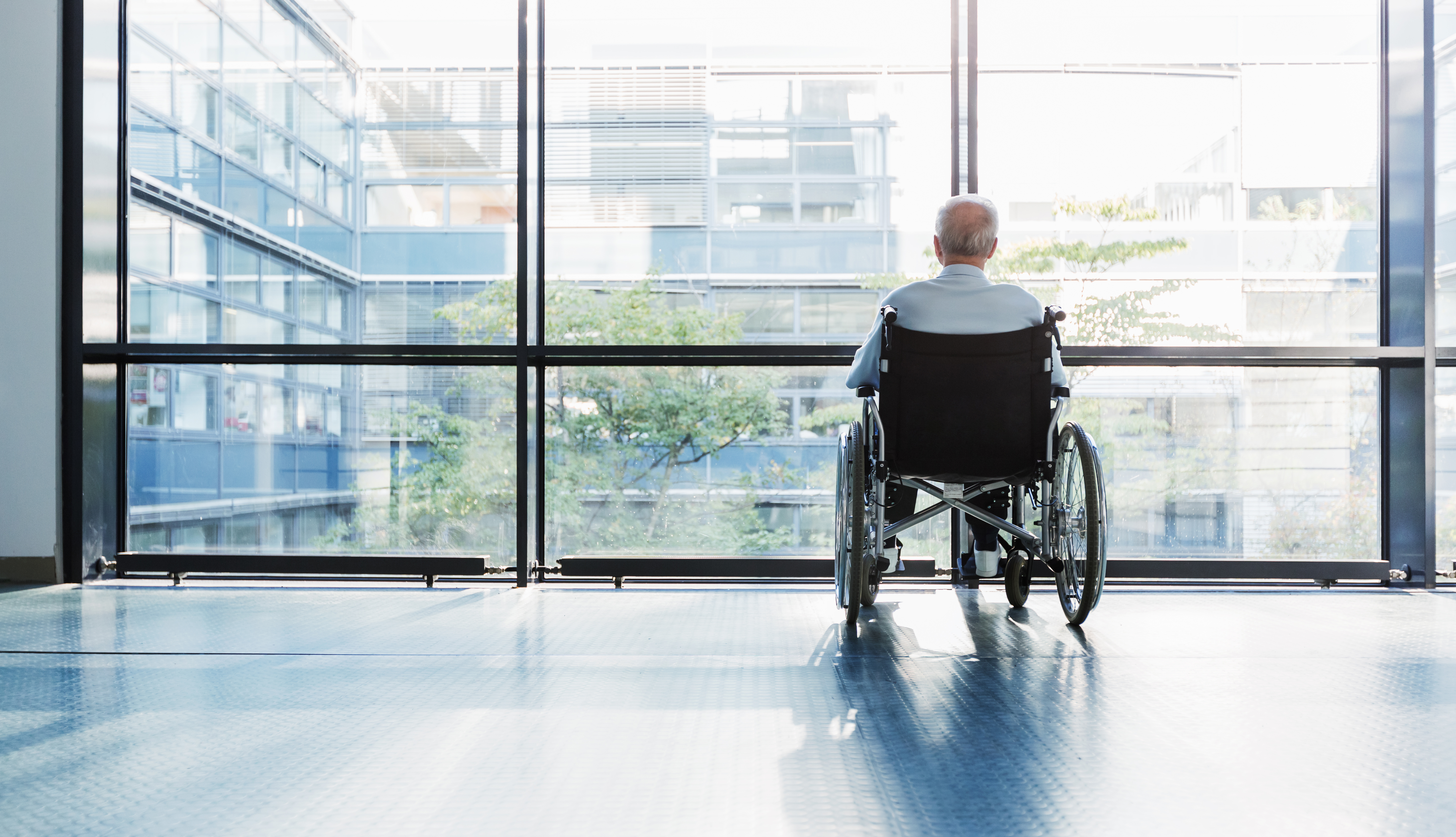 Senior Man in Wheelchair looking out of a window in a hospital corridor.