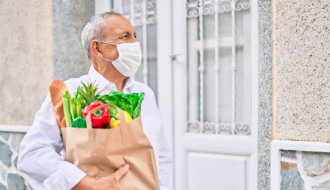 Older Caucasian man wearing a mask and holding a brown grocery bag full of fresh vegetables and baguette