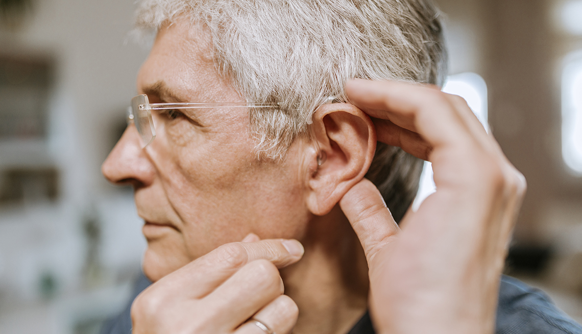 Hombre canoso sostiene su oreja preparándose para insertar un audífono con la otra mano