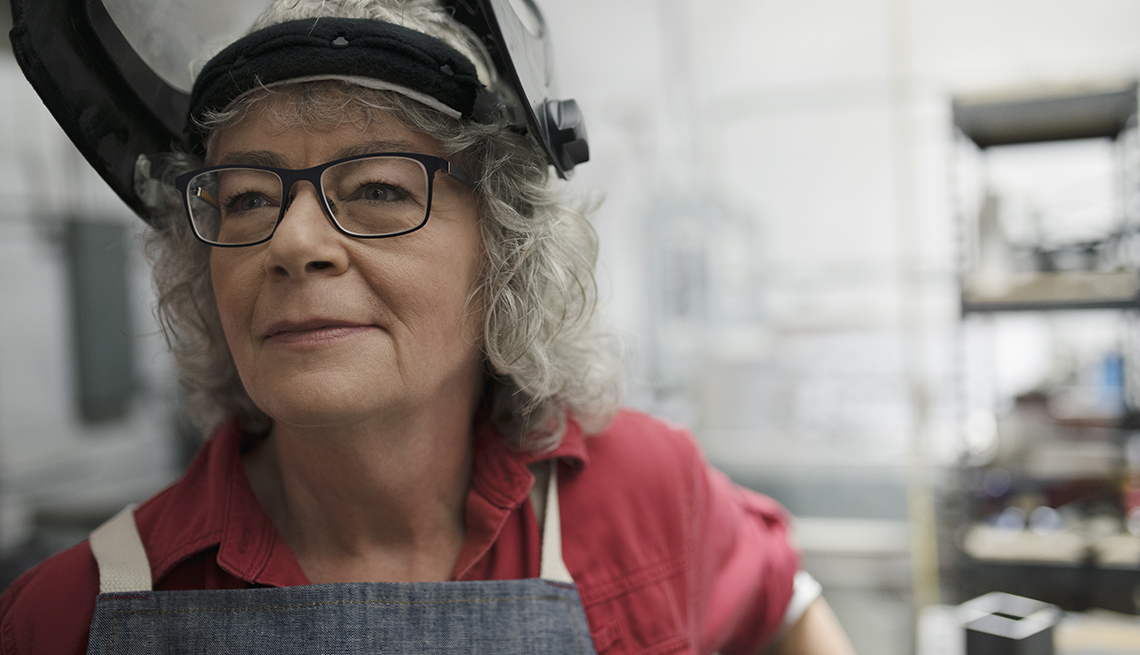 a female welder over age 50 is smiling at work with her face mask flipped up to show her face