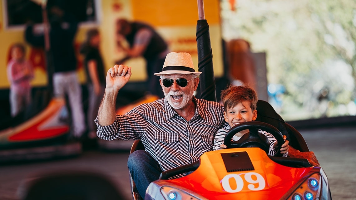 grandfather wearing sunglasses and plaid shirt with hat on and grandson riding in an orange with the number 9 on it go-cart smiling 