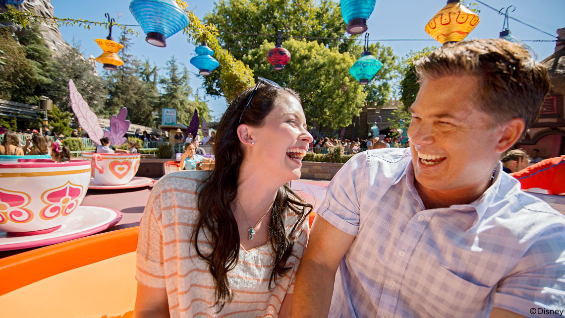 woman and man couple riding tea cups at theme park, both smiling and bright blue sky in background