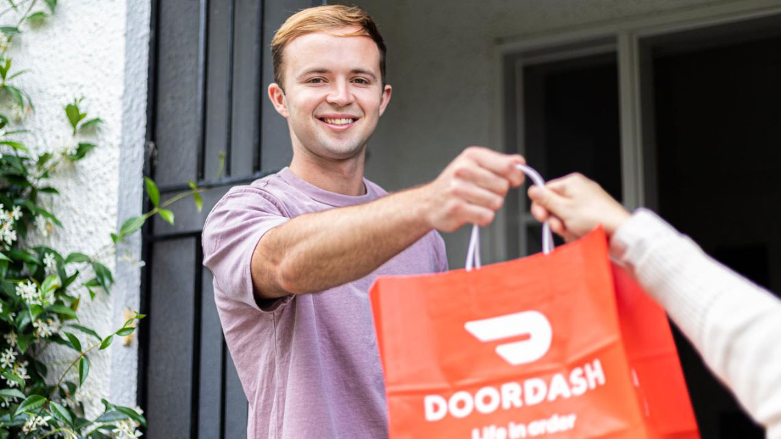 man with purple t-shirt smiling taking a doordash orange to-go bag from a delivery person