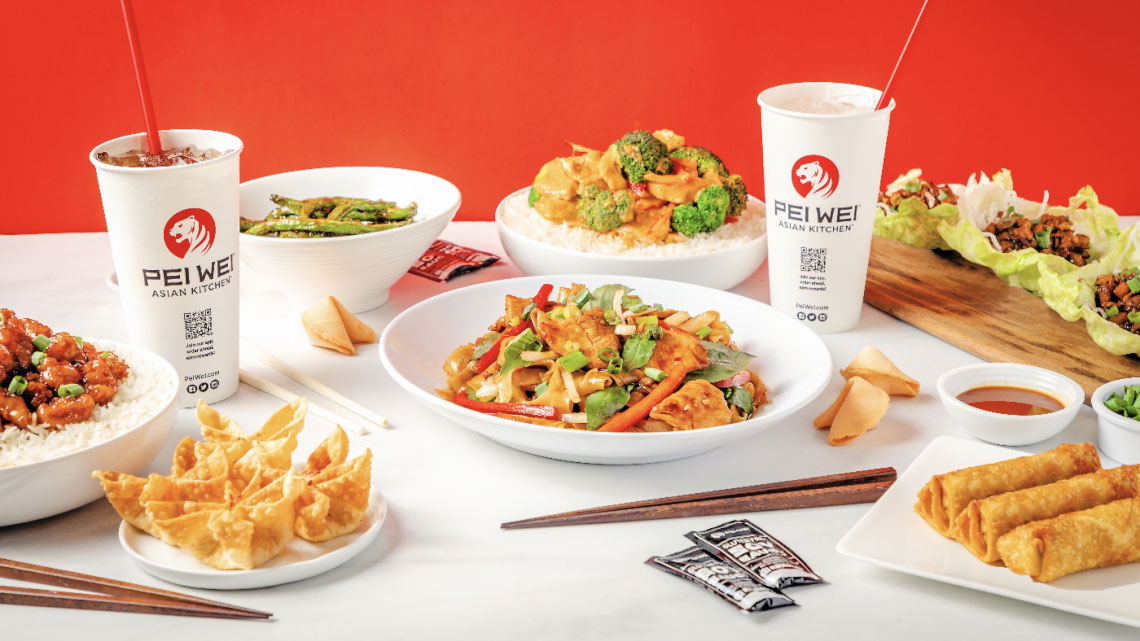 two soda cups with company logo in them surrounded by multiple Asian dishes including chopsticks, soy sauce packets, egg rolls, lettuce cups, rice and veggie bowls 