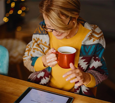 AARP member holding a cup of hot tea wearing a cozy sweater looking at AARP Rewards holiday resources on her tablet
