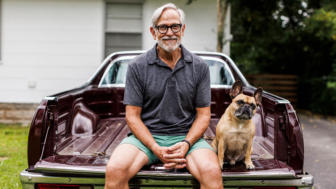 Portrait of smiling senior man sitting with dog on vintage car in driveway