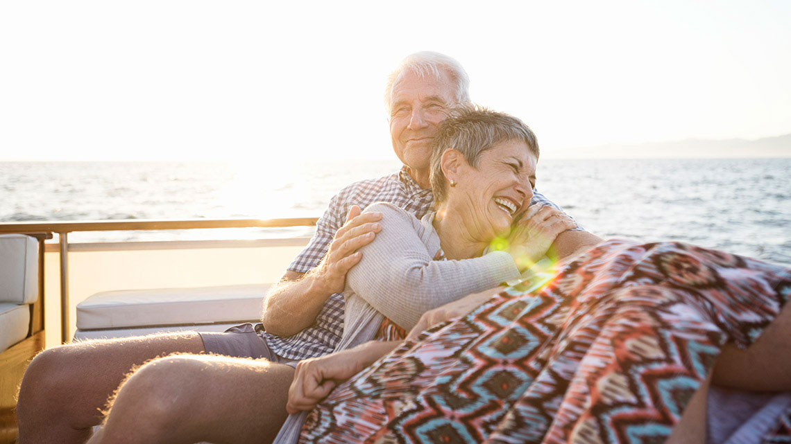 man and woman cozied up in a blanket sitting outside with beach in background both smiling, woman laughing