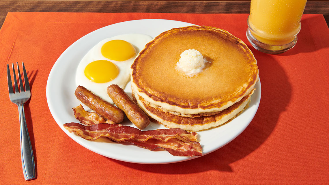 orange placemat with metal silver fork and glass of oj white plate with pancakes 2 eggs sausage bacon