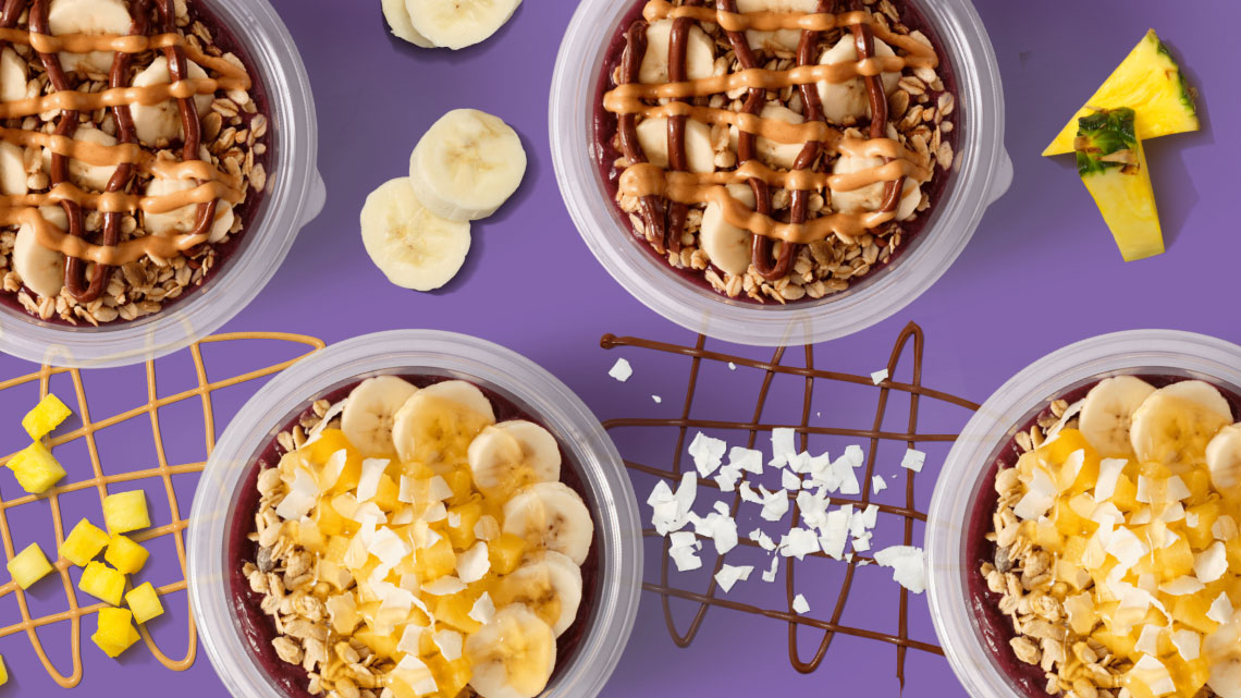 banana pieces, mango pieces, purple background, peanut butter, chocolate sauce, nuts, shaved coconut in bowls