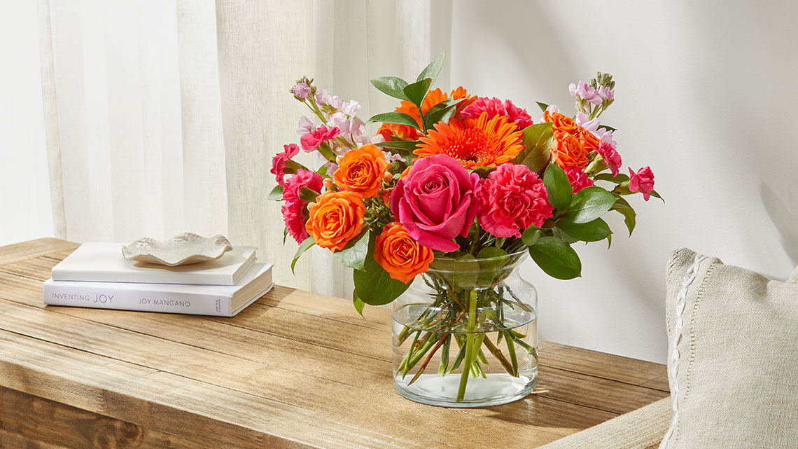 orange gerber daisies and roses with dark pink roses and carnations and other white, light purple flowers in a vase sitting on a wood table next to a linen colored chair