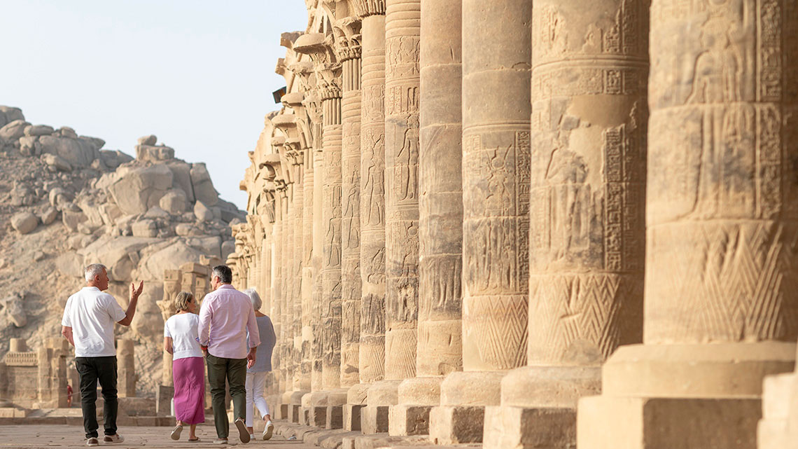 two men and two women walking outside next to ancient pillars, sand and rock terrain