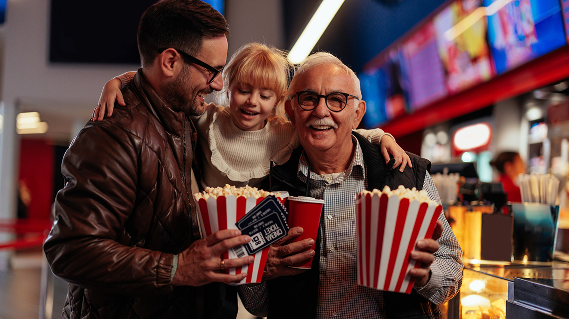 A grandfather, his son and his granddaughter are in the movie theater. They purchased tickets, soda, and popcorn and are headed to the movie hall to enjoy.