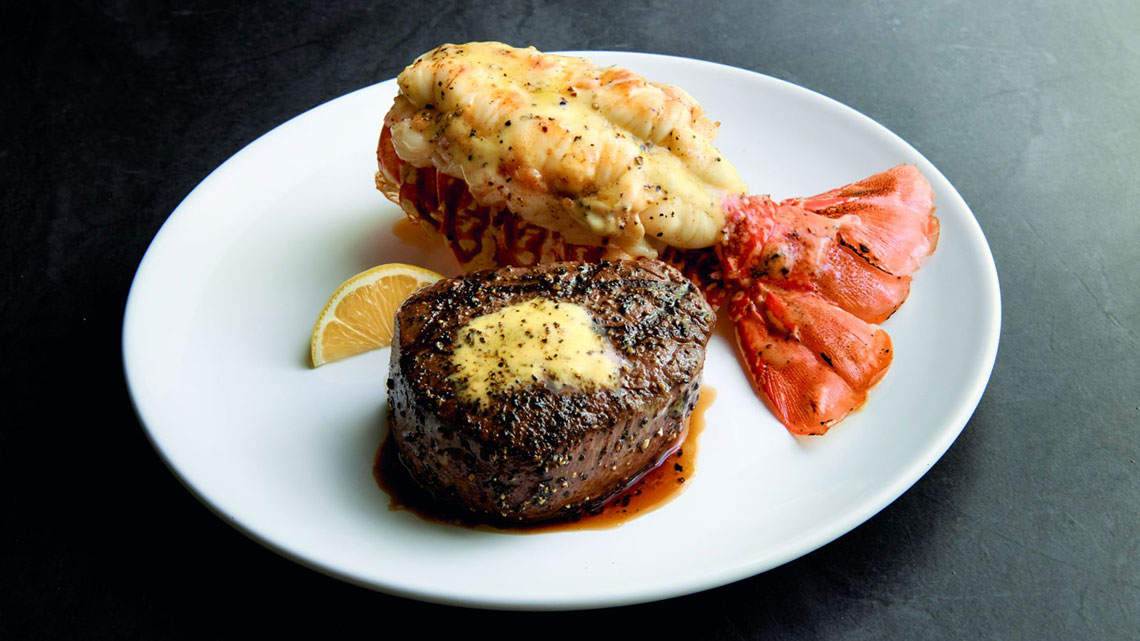 Butterflied lobster tail next to filet steak topped with butter, lemon wedge on side, all on top of white plate, dark gray background