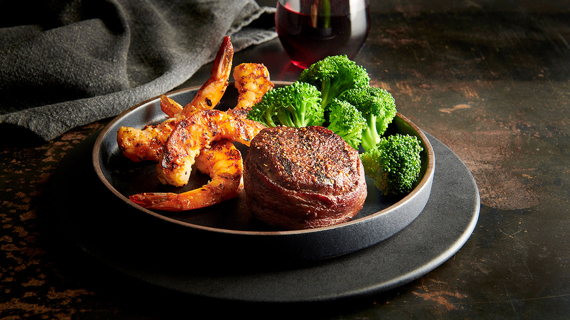 red glass wine next to a black plate of steak filet, shrimp, broccoli