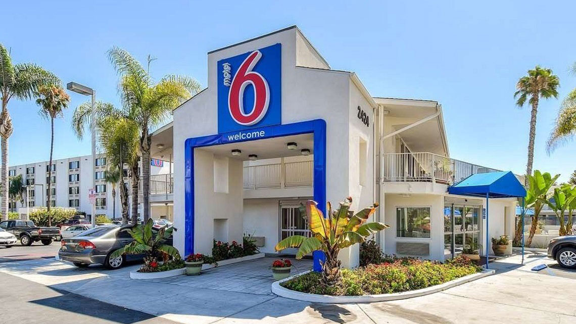 front of a Motel 6, large entrance way with blue and red logo on top, palm tree and other greenery around the entrance, cars in parking lot