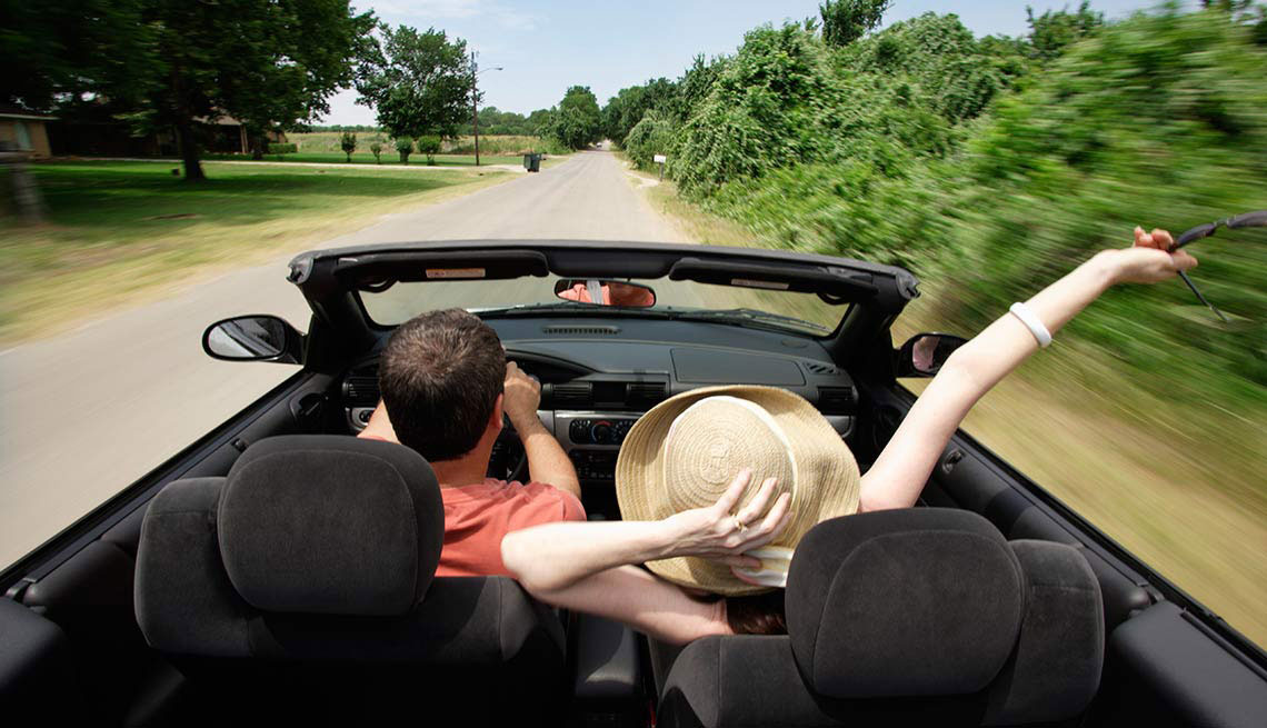 man and woman in top down convertible car driving on a country road, sunny day