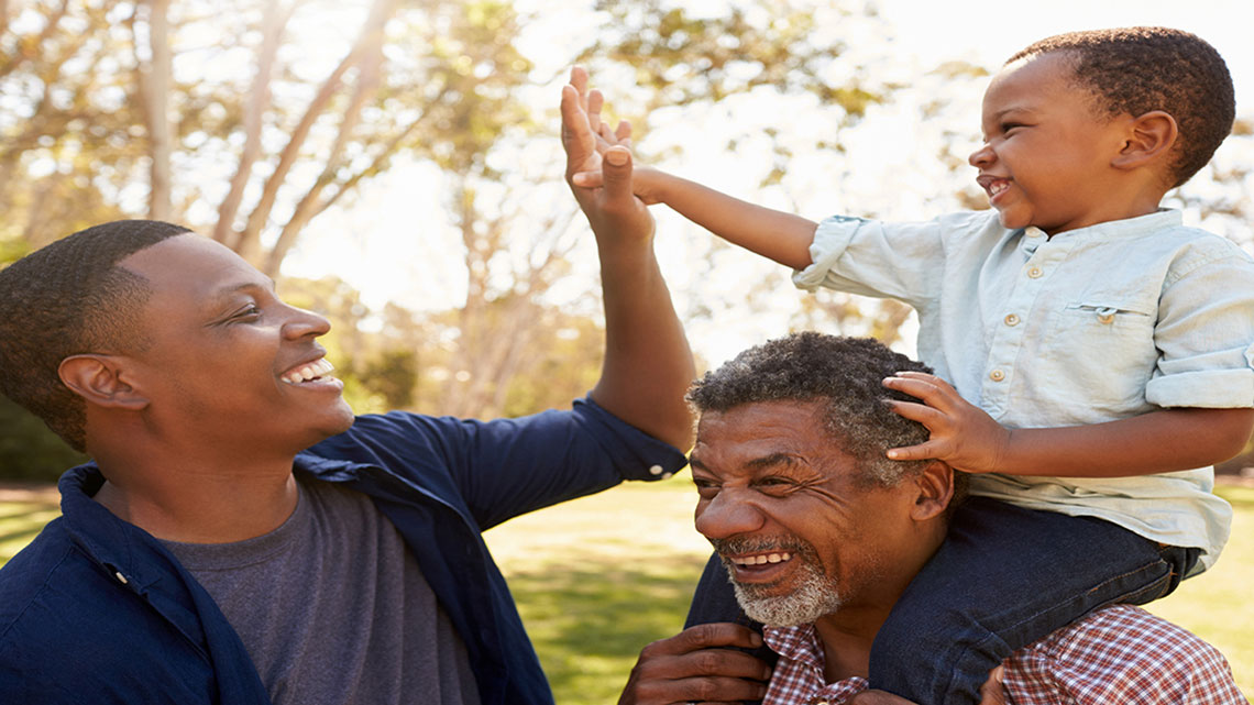 father high-fiving young son on shoulders of grandfather, all smiling, outside, sunny day