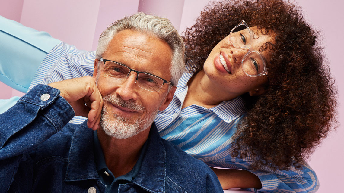 Man sitting in front of a girl laying posed behind him. both smiling, both wearing denim blue clothes, both wearing glasses, pink background