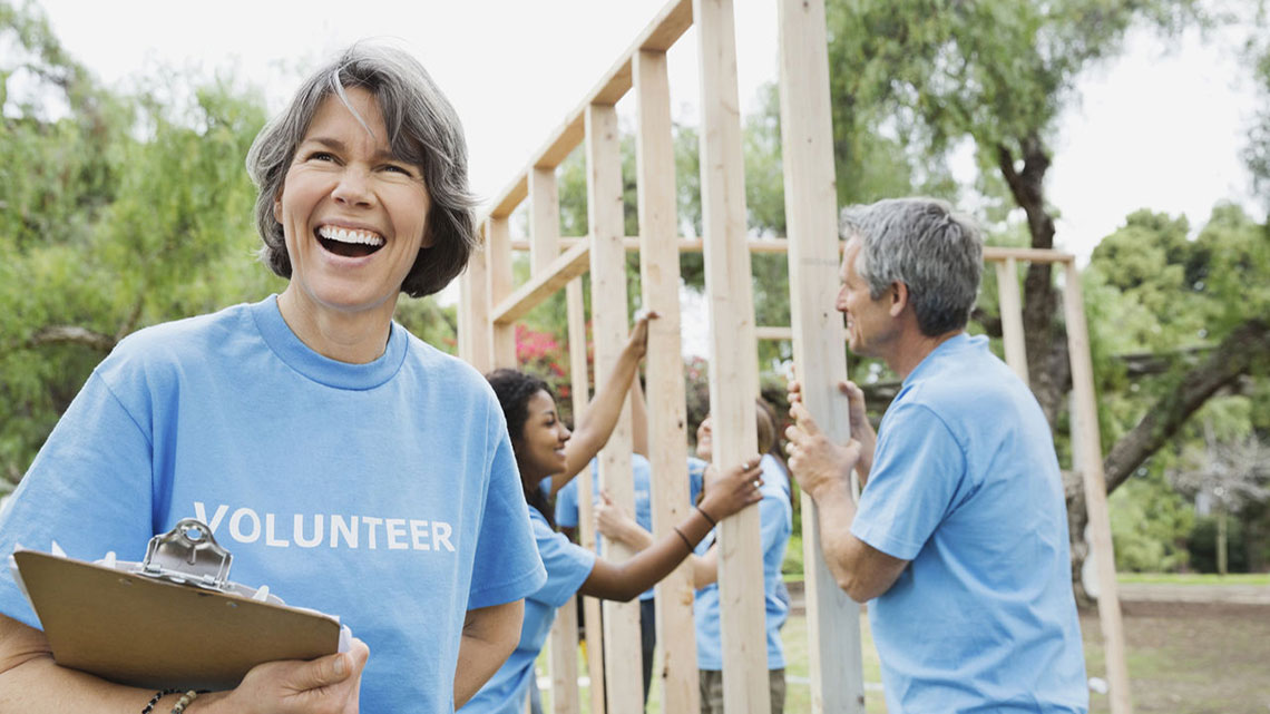 woman in blue tee shirt with the word volunteer in white lettering on the front, she is smiling holding a clipboard while a woman in background helps put up wood house framing