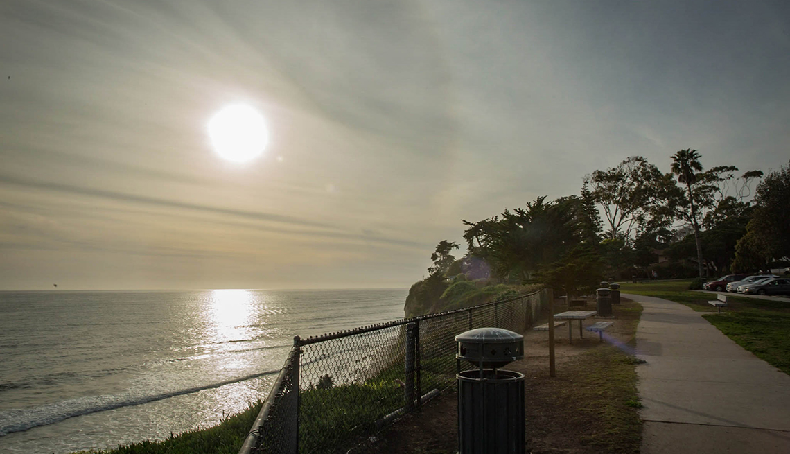 Park path, tables, fence and trees on bluff above the sea with sun going down over the water.