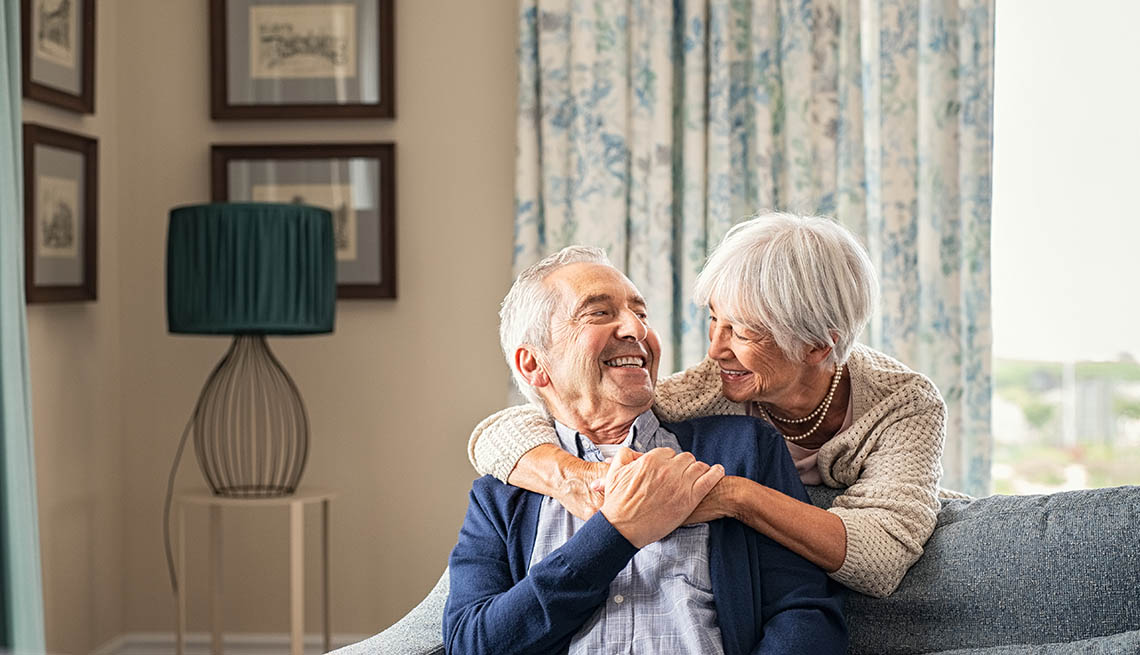 A woman embracing her husband at home while laughing together. Smiling wife hugging old man sitting on couch from behind. Joyful retired couple having fun at home while looking at each other with copy space. Love and unity concept.