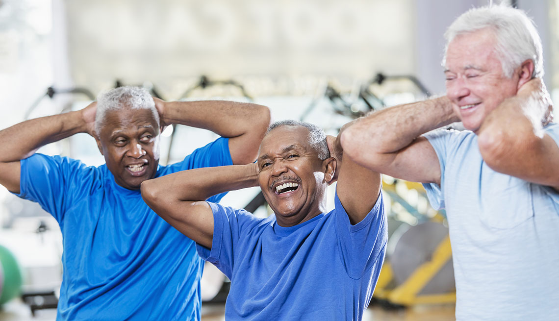 A group of three multi-ethnic senior men in their 60s having a good time as they exercise together. They are sitting on fitness balls, hands behind their heads. The focus is on the African-American man in the middle who is laughing.