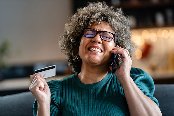 Shocked woman with curly hair and glasses is holding a credit card while talking on her phone, looking concerned 