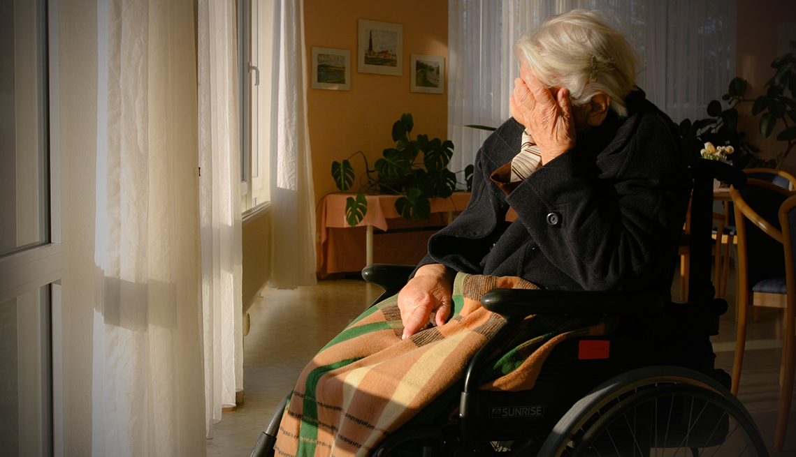 A man sitting in a wheelchair in a nursing home with his head in his hand, looking sad