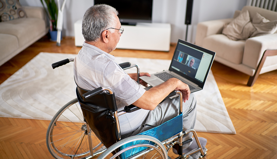 Hombre en su sala sentado en una silla de ruedas usando una computadora portátil para una cita de telesalud con su médico.
